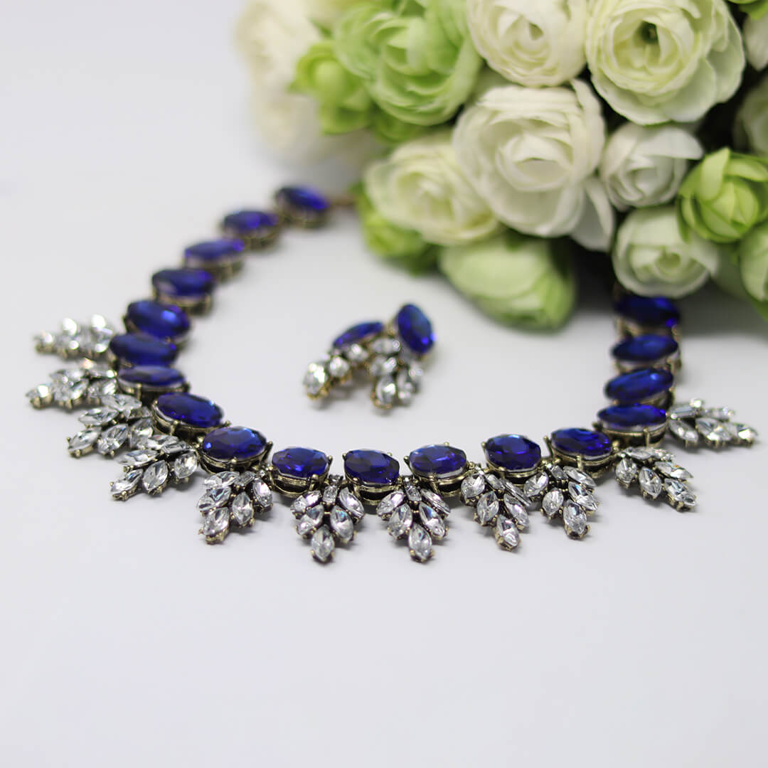 A jewelry set featuring a silver-toned necklace and matching earrings with deep blue oval stones and clear crystal leaf motifs, styled with white and light green roses on a white background.