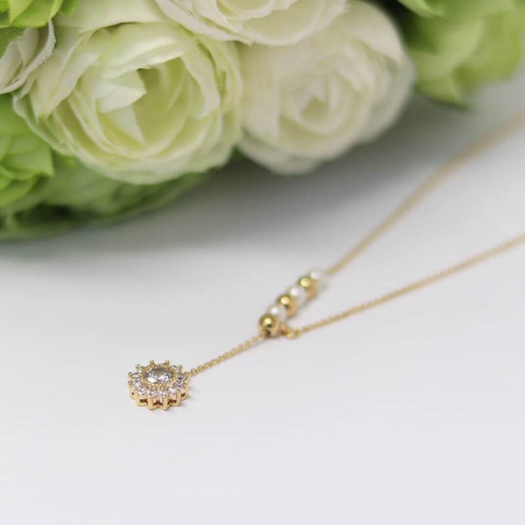 A delicate gold necklace with a small sunburst pendant set with clear stones, resting on a white surface with soft-focus white roses in the background.