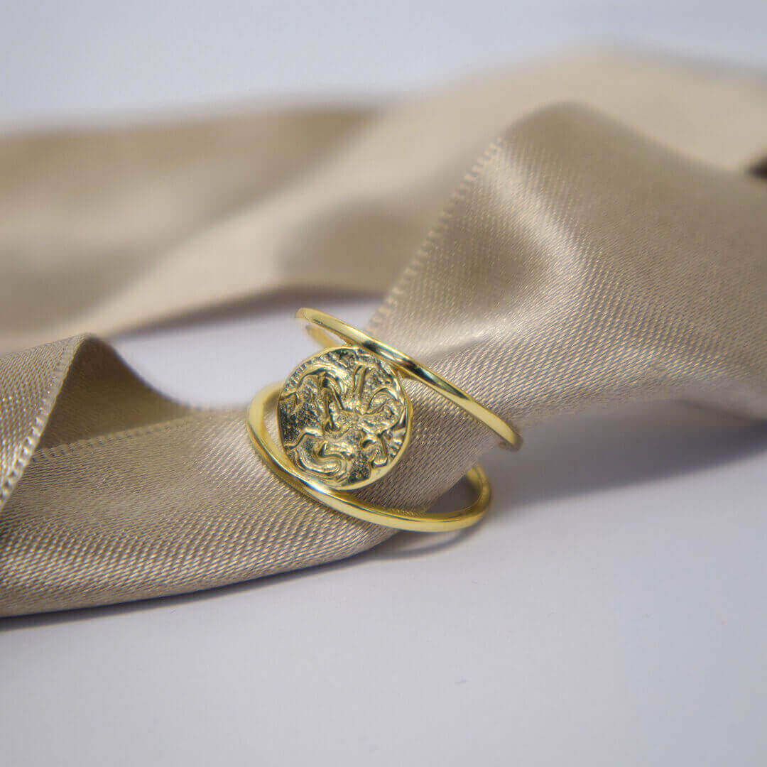 Gold ring with intricate design on a beige fabric background-Belle de Soiree
