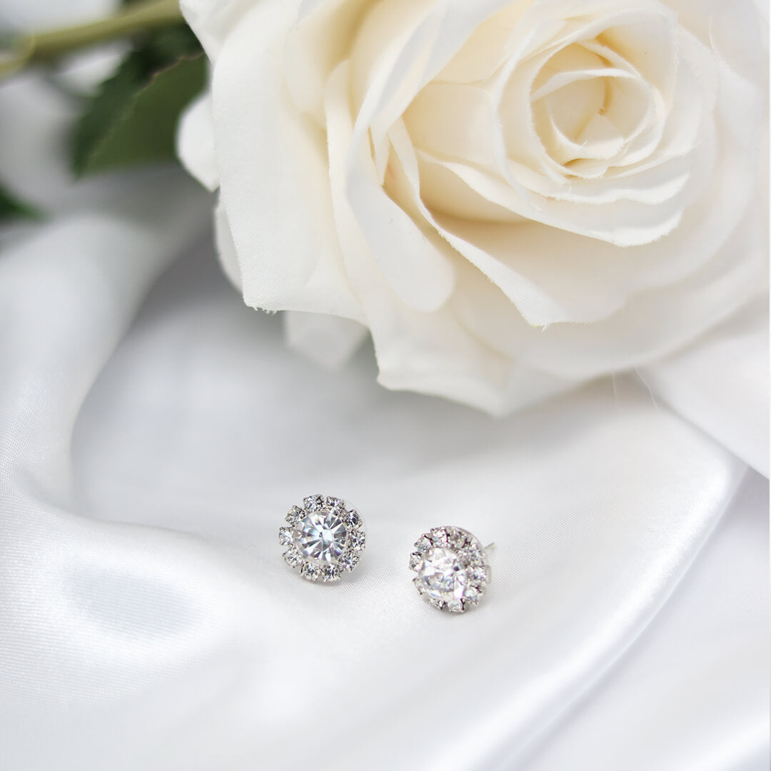 A pair of round stud earrings with sparkling clear gemstones in silver-toned settings, displayed on white satin fabric next to a large white rose.