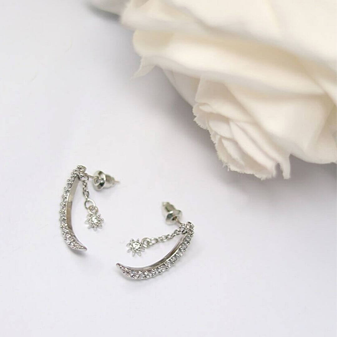 A pair of silver crescent moon earrings adorned with clear rhinestones and small dangling star charms, displayed on a white surface next to a white rose.