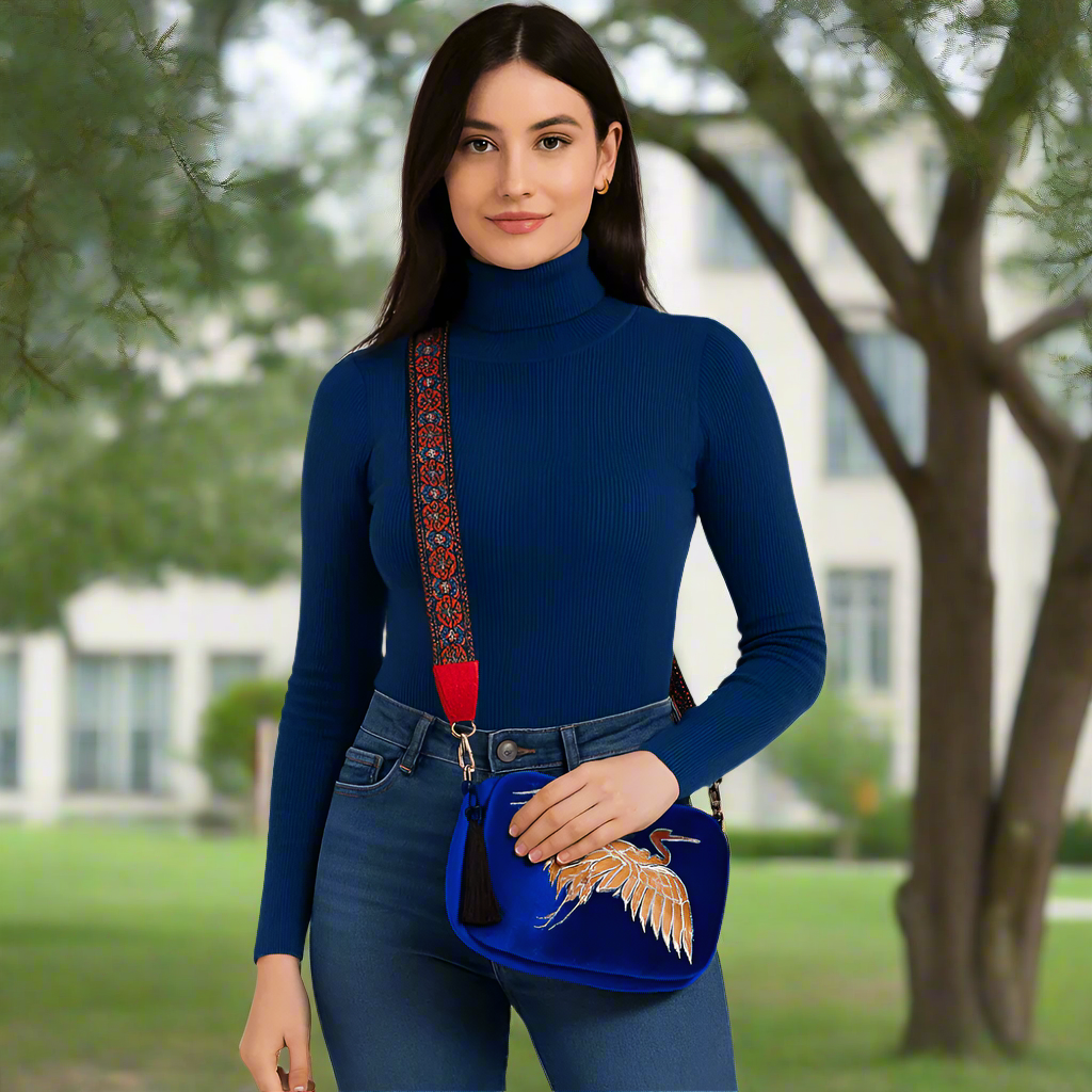 Woman wearing a blue turtleneck sweater and jeans, holding a blue handbag with a decorative strap.